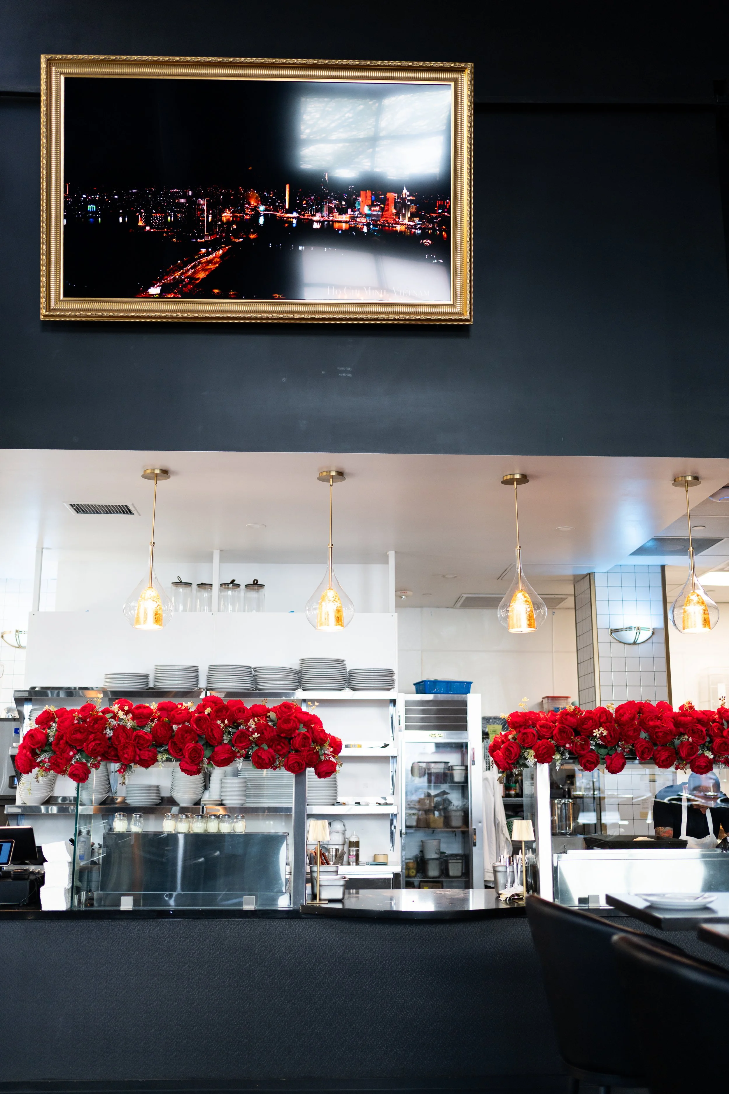 Restaurant interior featuring a black wall with a framed cityscape picture, white kitchen with shelves of plates, two bouquets of red roses, and hanging pendant lights.