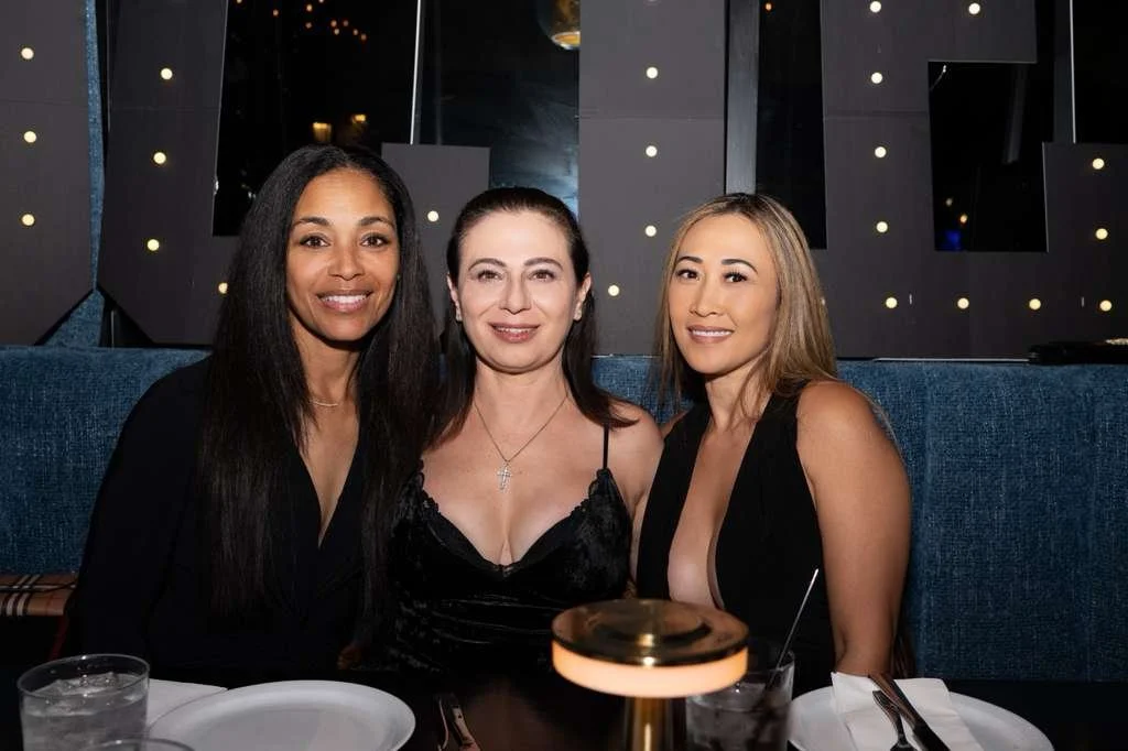 Three women sitting together at a restaurant table, dressed in black, smiling at the camera, with a dark interior background and table settings including glasses and plates.