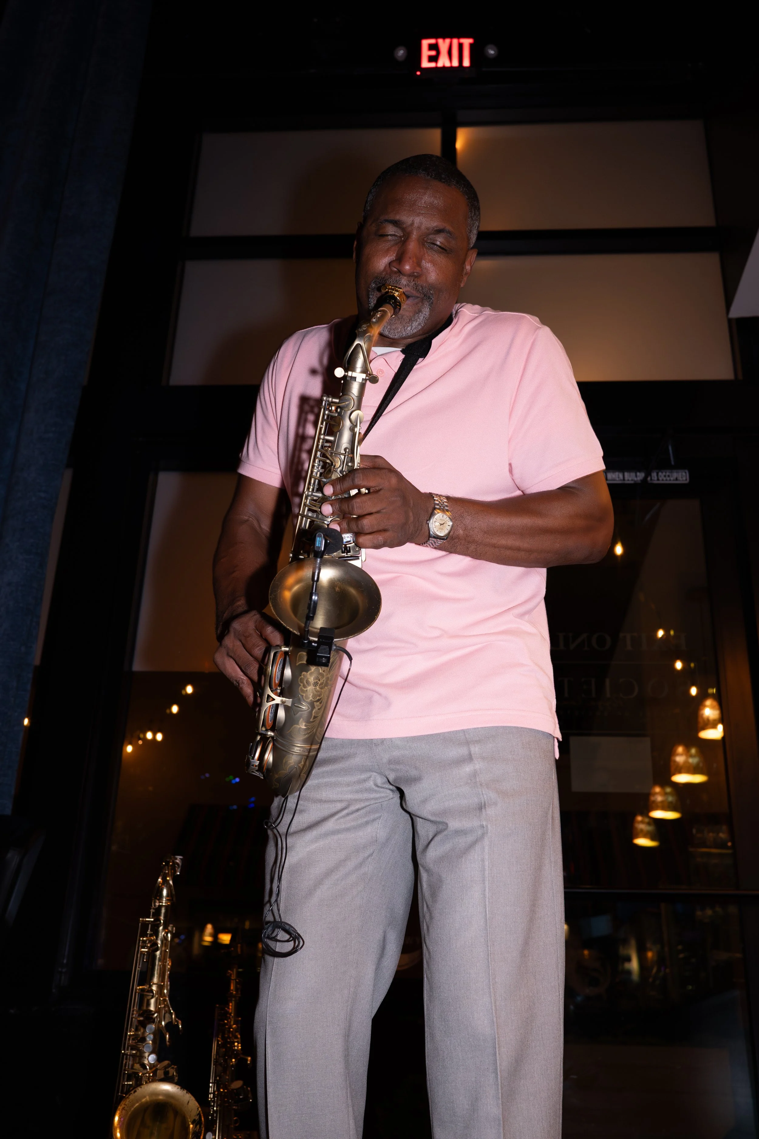 A man playing a saxophone at night, wearing a pink shirt and gray pants, with an exit sign illuminated above him.