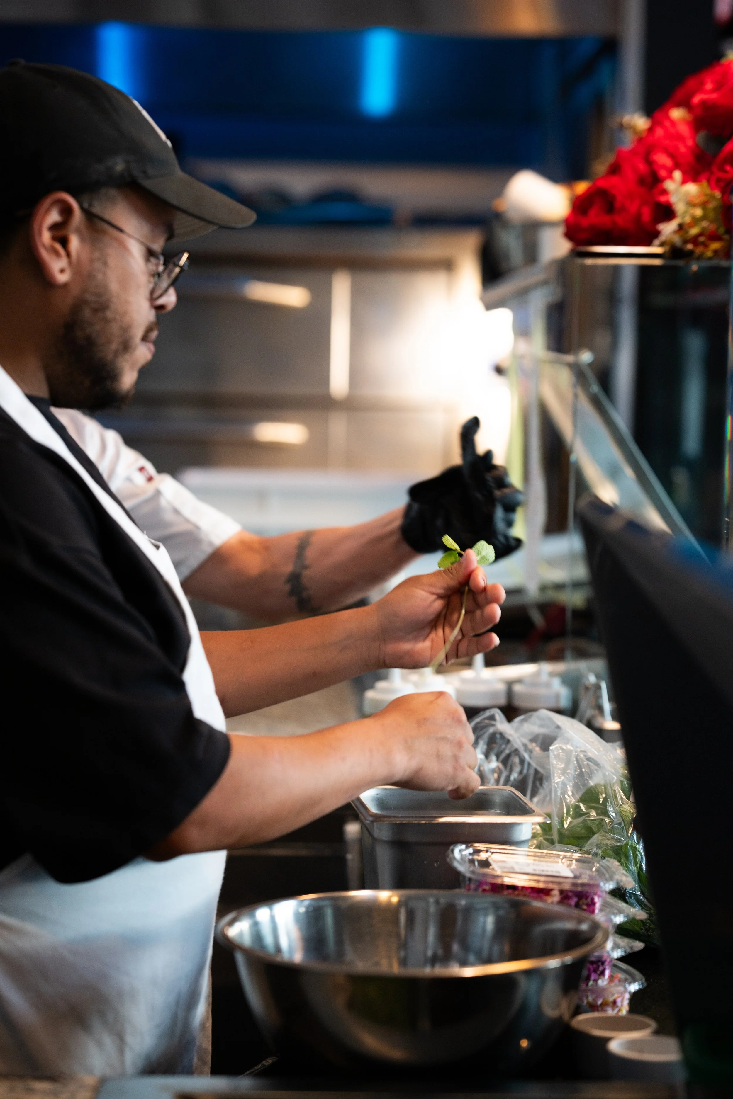 A chef preparing food in a professional kitchen, wearing glasses, a black cap, and a white apron, with various ingredients and kitchen tools on the counter.
