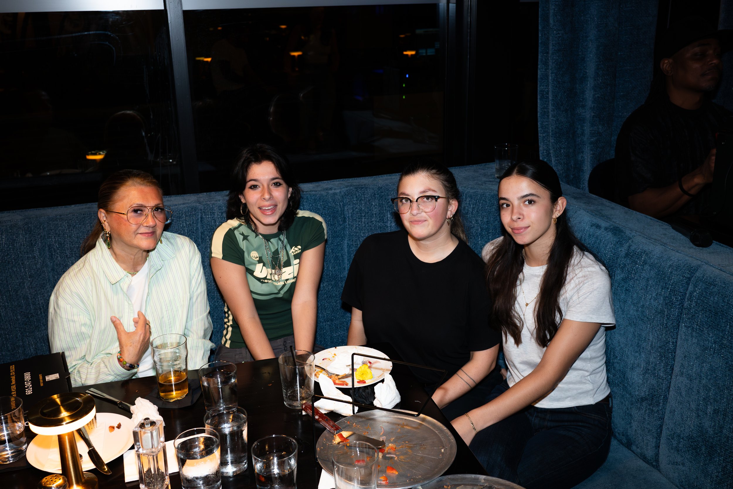 A group of four women sitting at a restaurant table after eating, with empty and partially eaten plates, glasses, and utensils. They are smiling and posing for the photo, with a blue booth and dark background behind them.