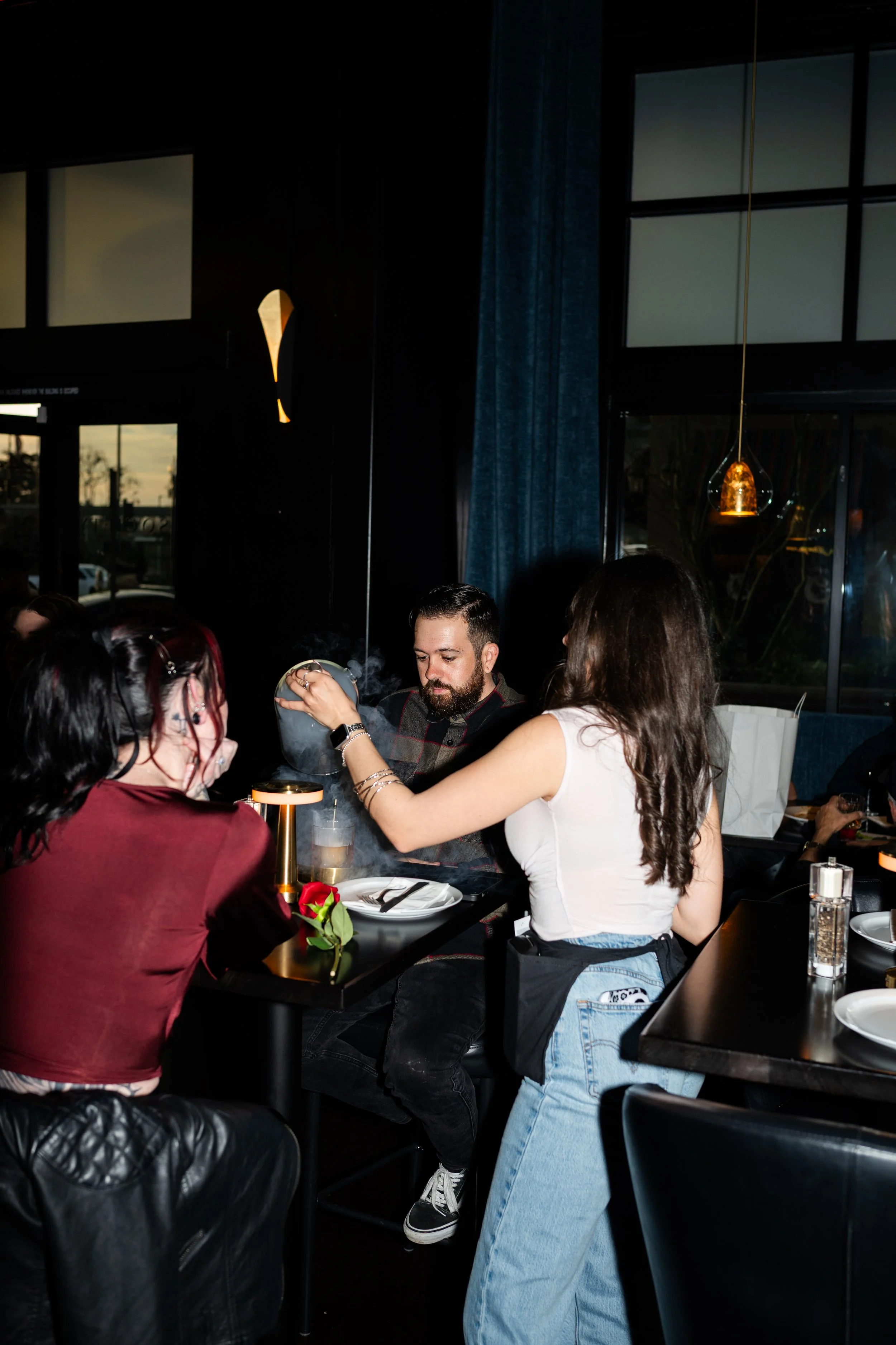 A restaurant scene with four people gathered around a table. One woman is helping a man with a steaming dish. The restaurant has dark walls, large windows, and modern pendant lighting.