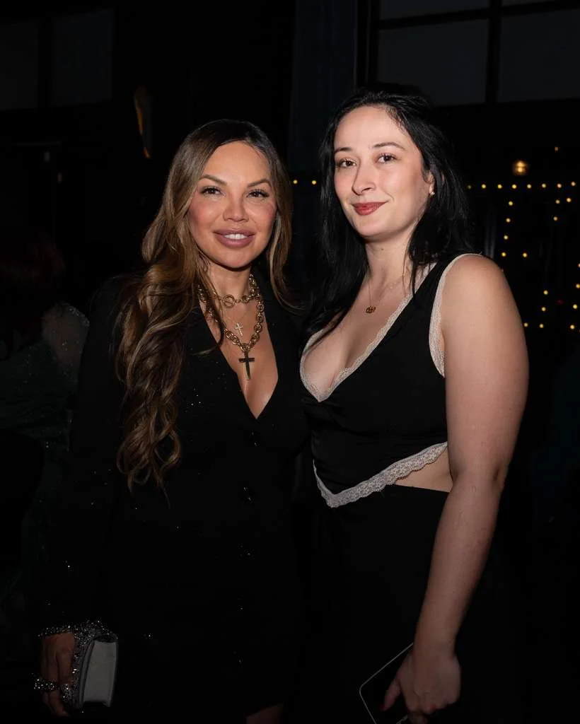 Two women smiling and posing together at an indoor event, dressed in stylish outfits with dark backgrounds and string lights in the distance.