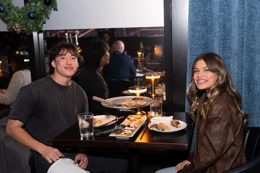 A young man and woman sitting at a restaurant table with pizza and drinks, smiling. Christmas tree decoration visible in the background.