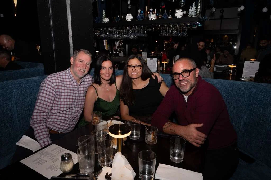 Four people sitting at a restaurant table smiling, with drinks and menus, in a dimly lit, festive environment with holiday decorations.