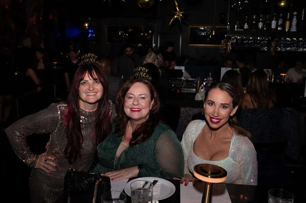 Three women at a New Year's celebration in a dimly lit restaurant or bar, wearing party hats that say "Happy New Year," smiling, with other people in the background.