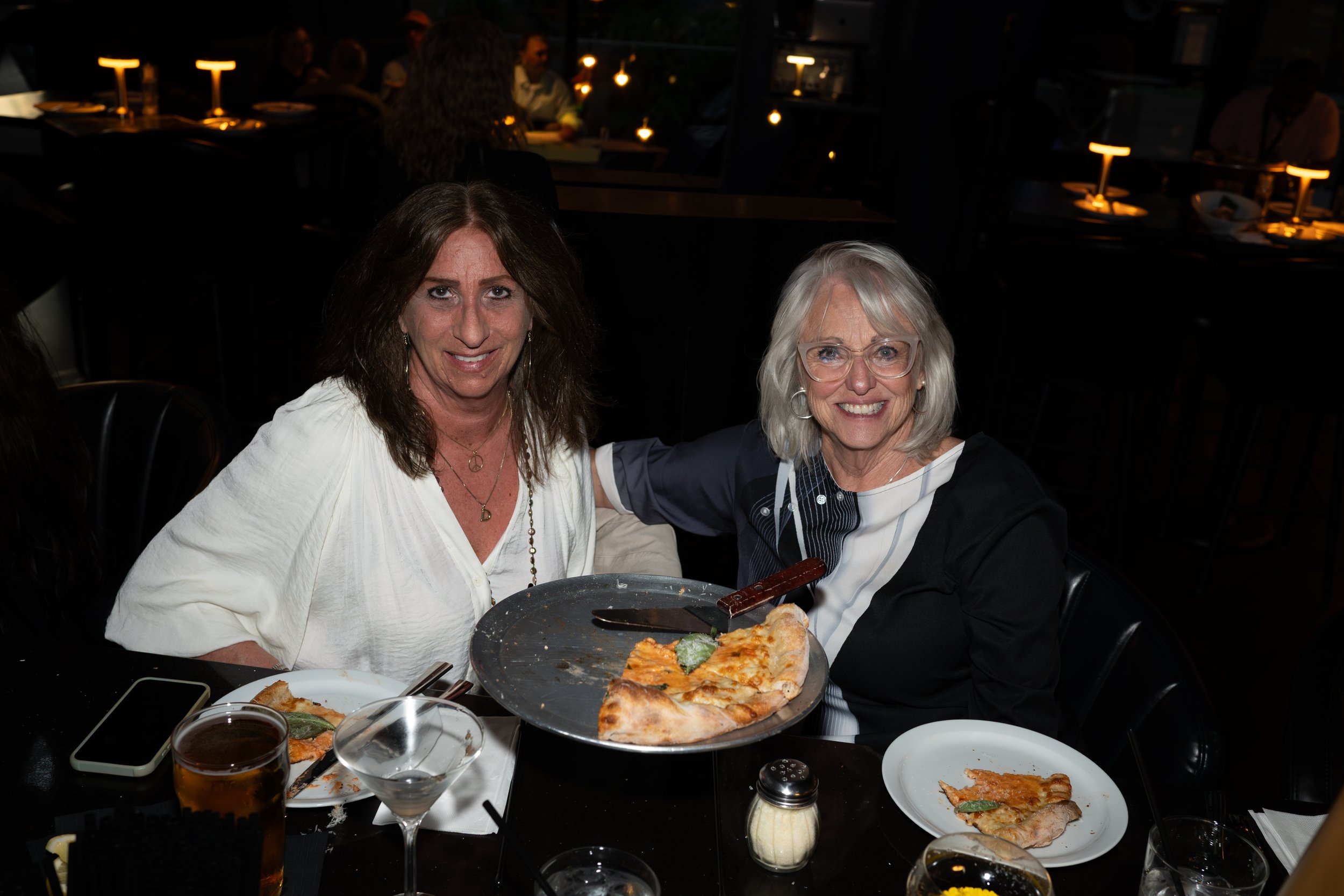Two women sitting at a dinner table in a dimly lit restaurant, smiling at the camera. The table has pizza, drinks, and dinnerware. The background shows other patrons and warm lighting.