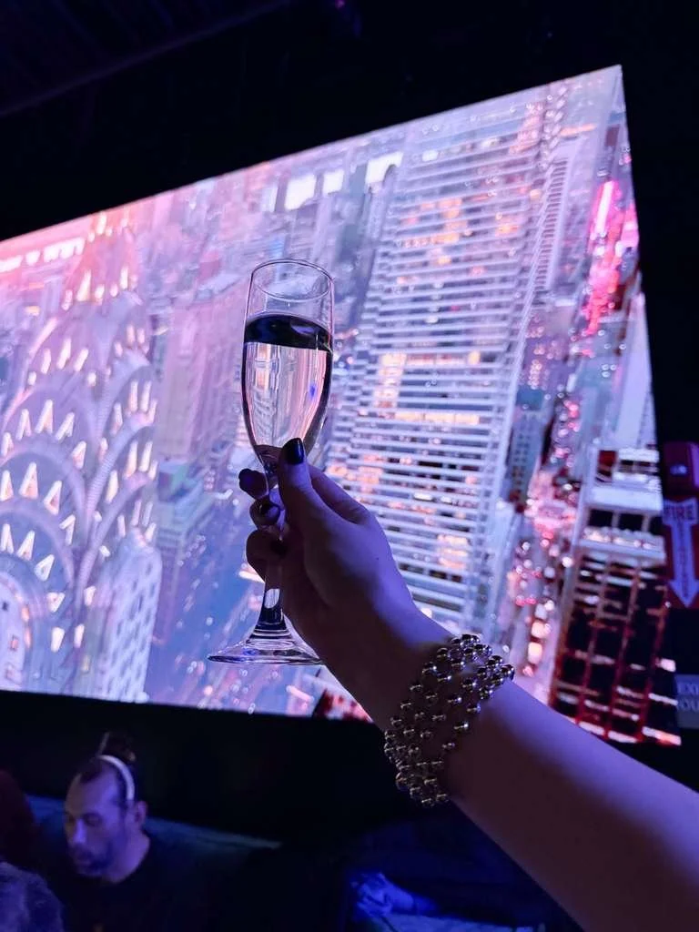 A person holding a glass of champagne in front of a large digital screen displaying a cityscape with tall buildings, likely in New York City.