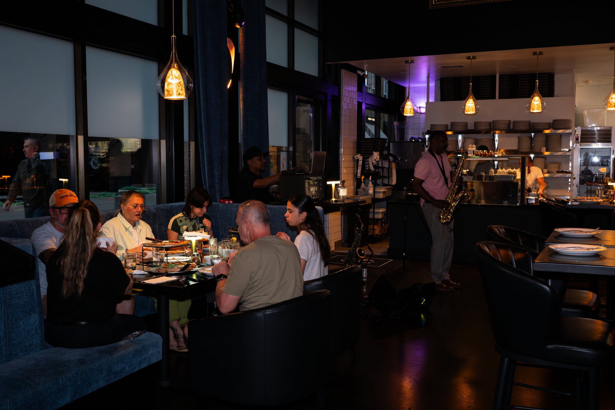 Interior of a restaurant with hanging pendant lights, a bar area, and a live musician playing saxophone. Customers are dining at tables.
