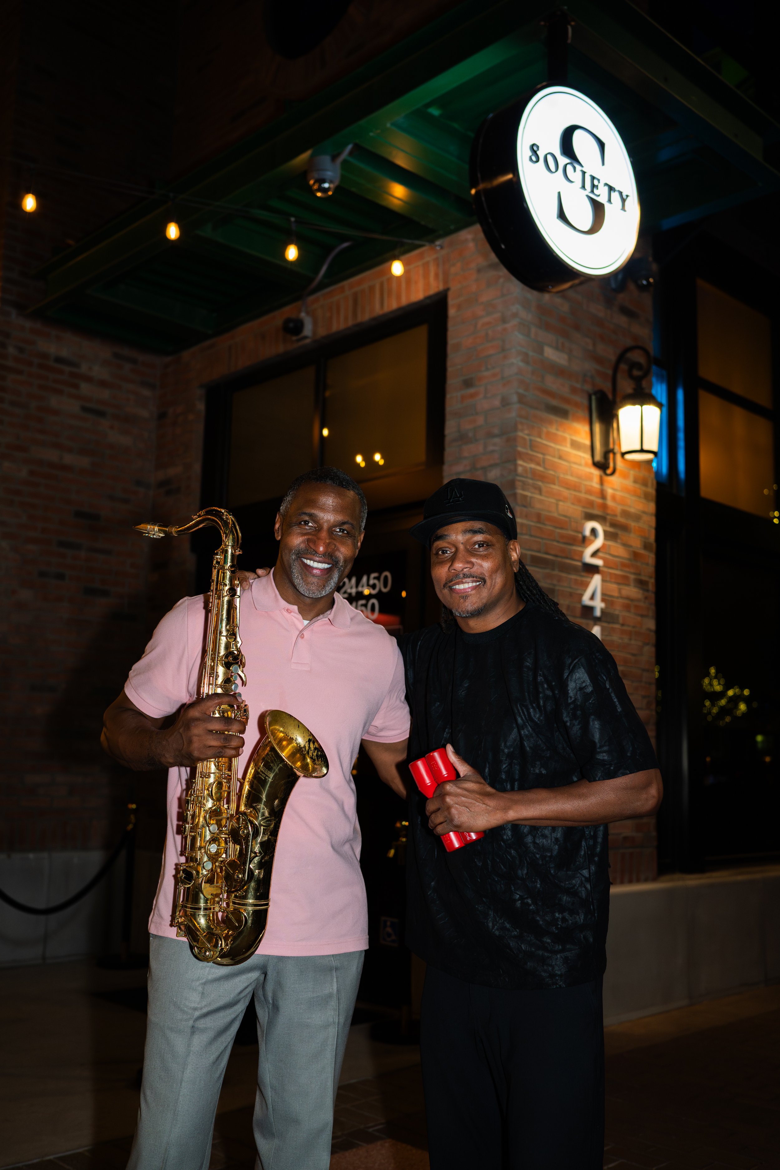 Two men standing outside a brick building at night, smiling. One man is holding a saxophone, and the other is holding a red object. There is a glowing sign above them that says 'Society' with a stylized 'S' logo. The scene is lit with warm outdoor li