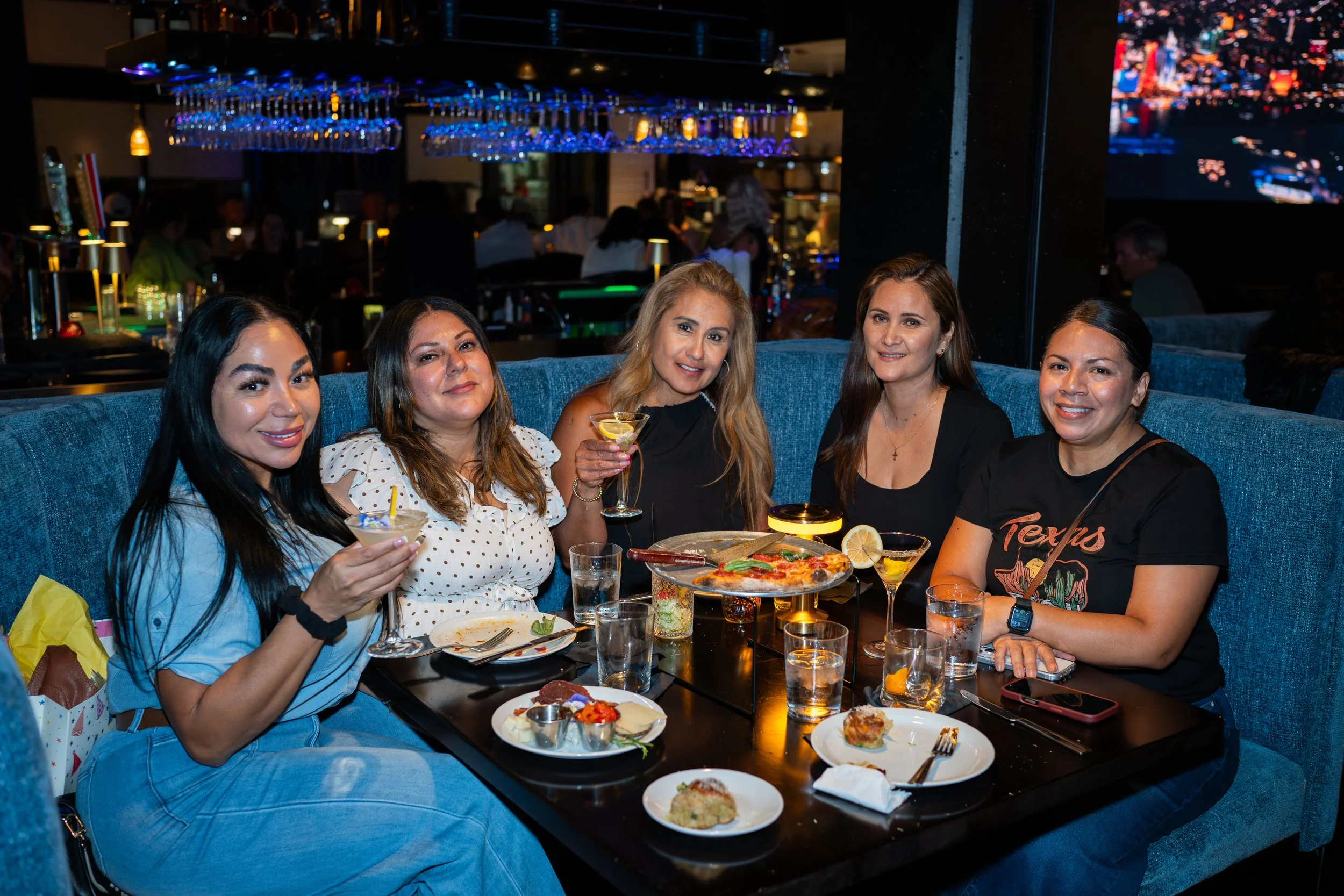 Group of five women sitting at a restaurant table enjoying drinks and food, with a background of a dimly lit bar area and city lights outside.
