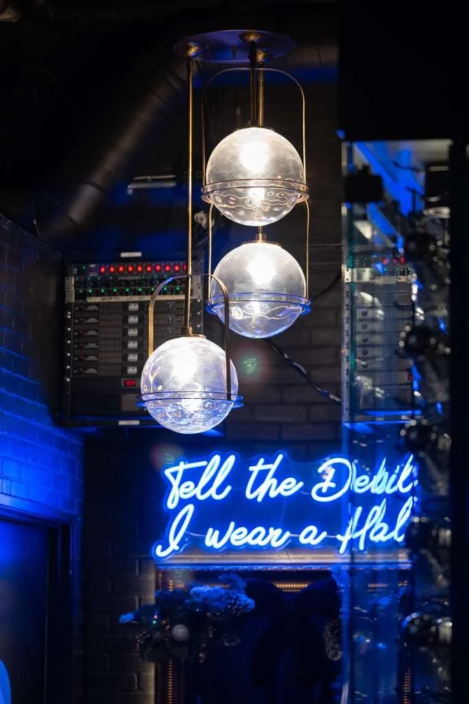 Neon sign that reads "Tell the Devil I wear a Halo" illuminated in blue, with a modern hanging light fixture with three frosted globes hanging above. The background features a dark brick wall.