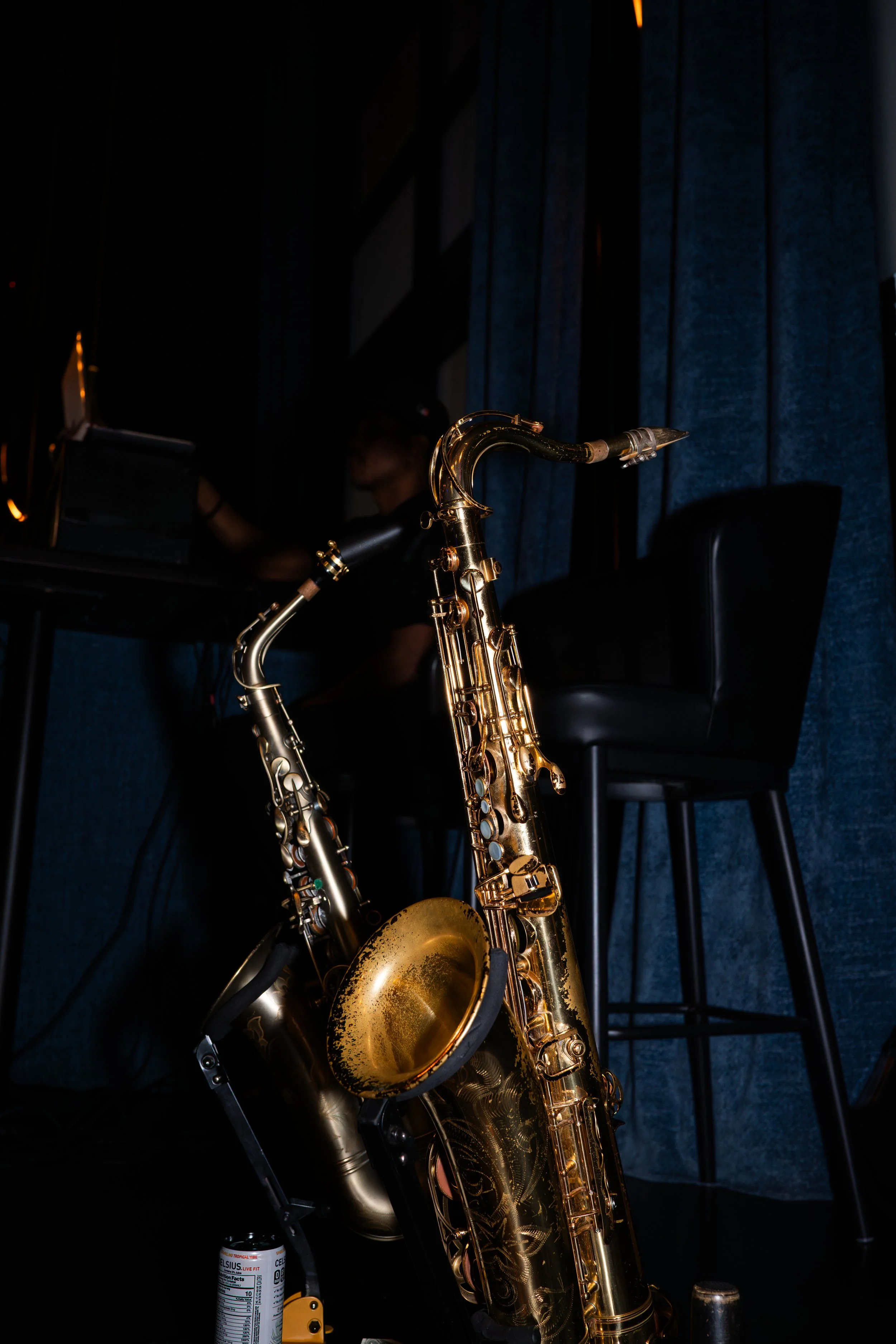 A gold-colored saxophone in the foreground with a dark stage background and a person at a keyboard in the background.
