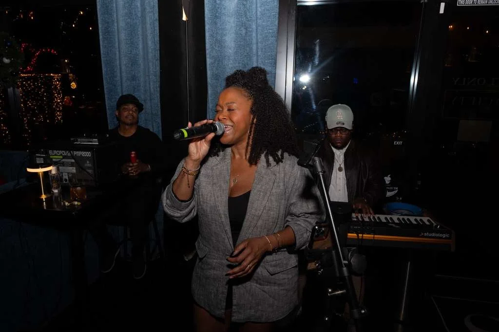 A woman singing with a microphone at a bar or lounge, with two musicians playing instruments in the background at night.