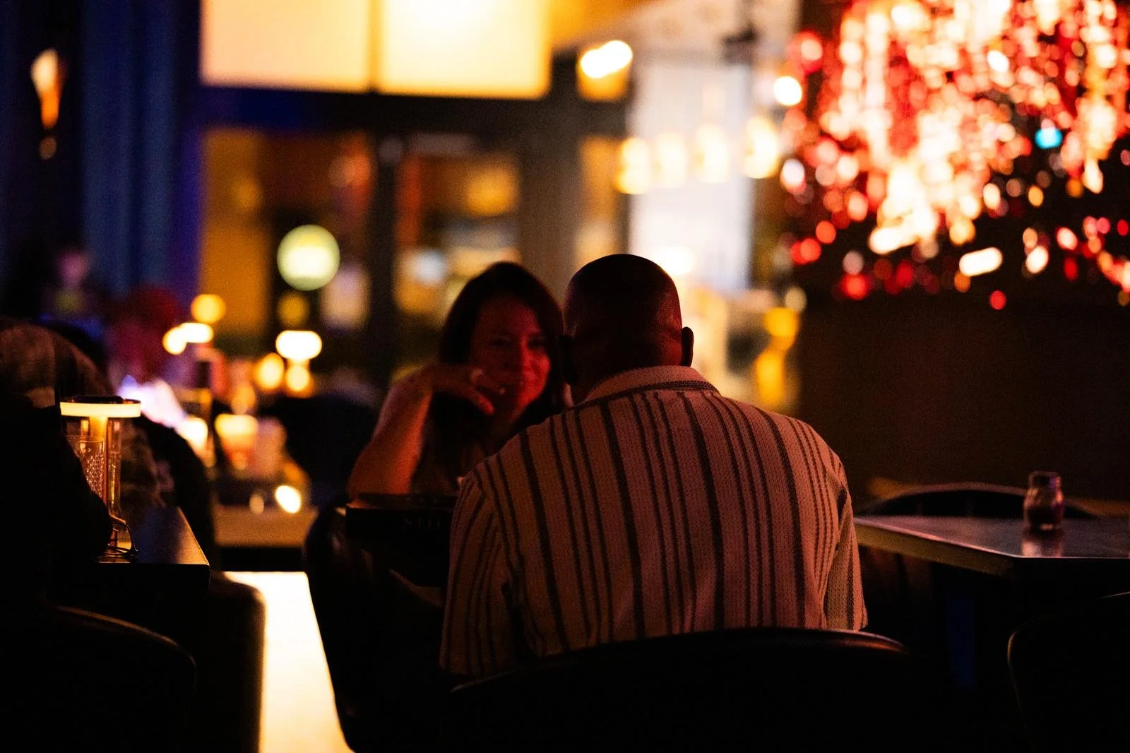 A man and woman sitting at a dimly lit table in a bar or restaurant, with neon and colorful lights and blurred background.
