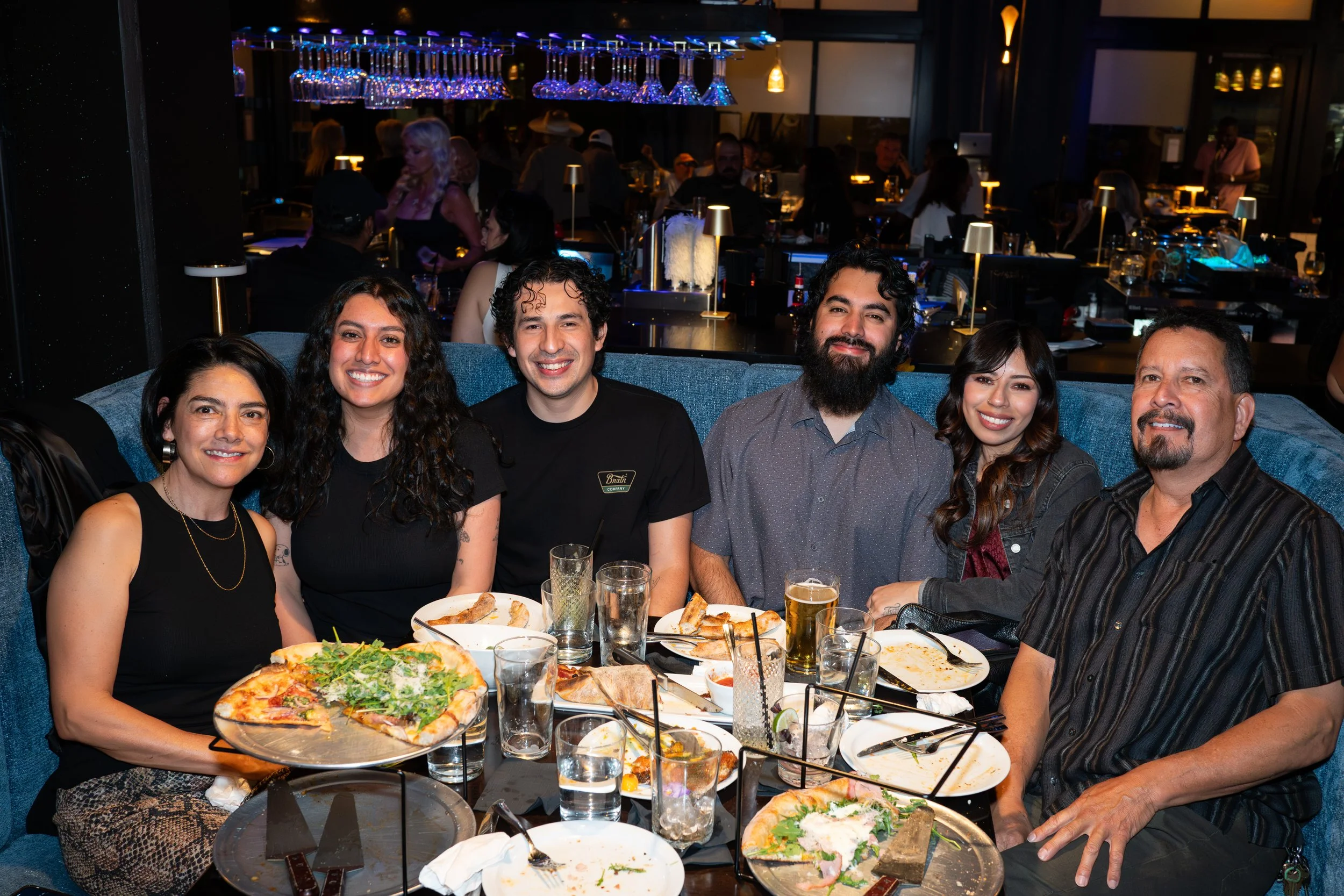 A group of six people sitting at a restaurant table, smiling at the camera, with pizza, drinks, and plates on the table. The restaurant has dim lighting and an upscale ambiance, with other diners visible in the background.