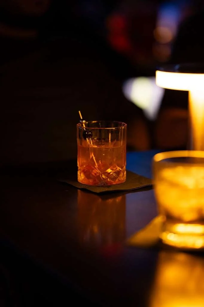 A glass of orange-colored cocktail with a cherry garnish on a dark table lit by warm ambient light.
