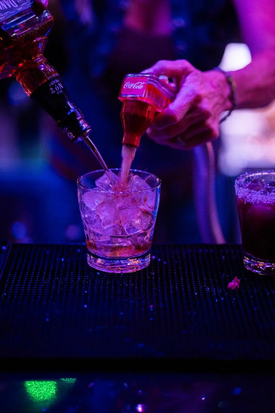 Bottled Coca-Cola being poured into a glass with ice at a bar, with neon lighting and another drink nearby.