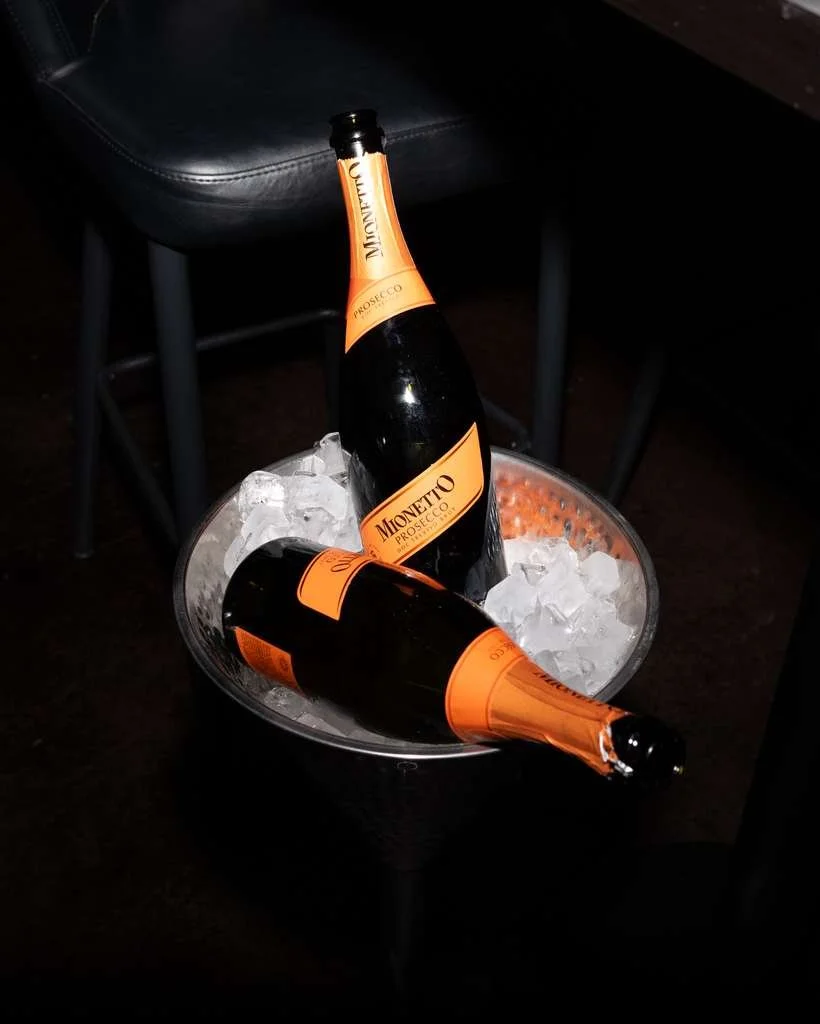 Two bottles of Monetio Prosecco in an ice bucket filled with ice cubes, placed on a dark table.