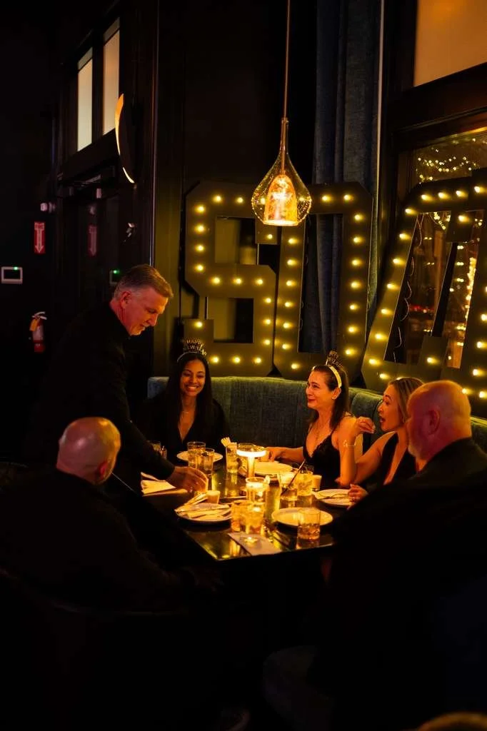 A birthday celebration in a dimly lit restaurant with six people sitting around a table with drinks and snacks. A woman wearing a birthday crown sits in the center, smiling. A man is serving drinks. Large illuminated letters display a number behind t