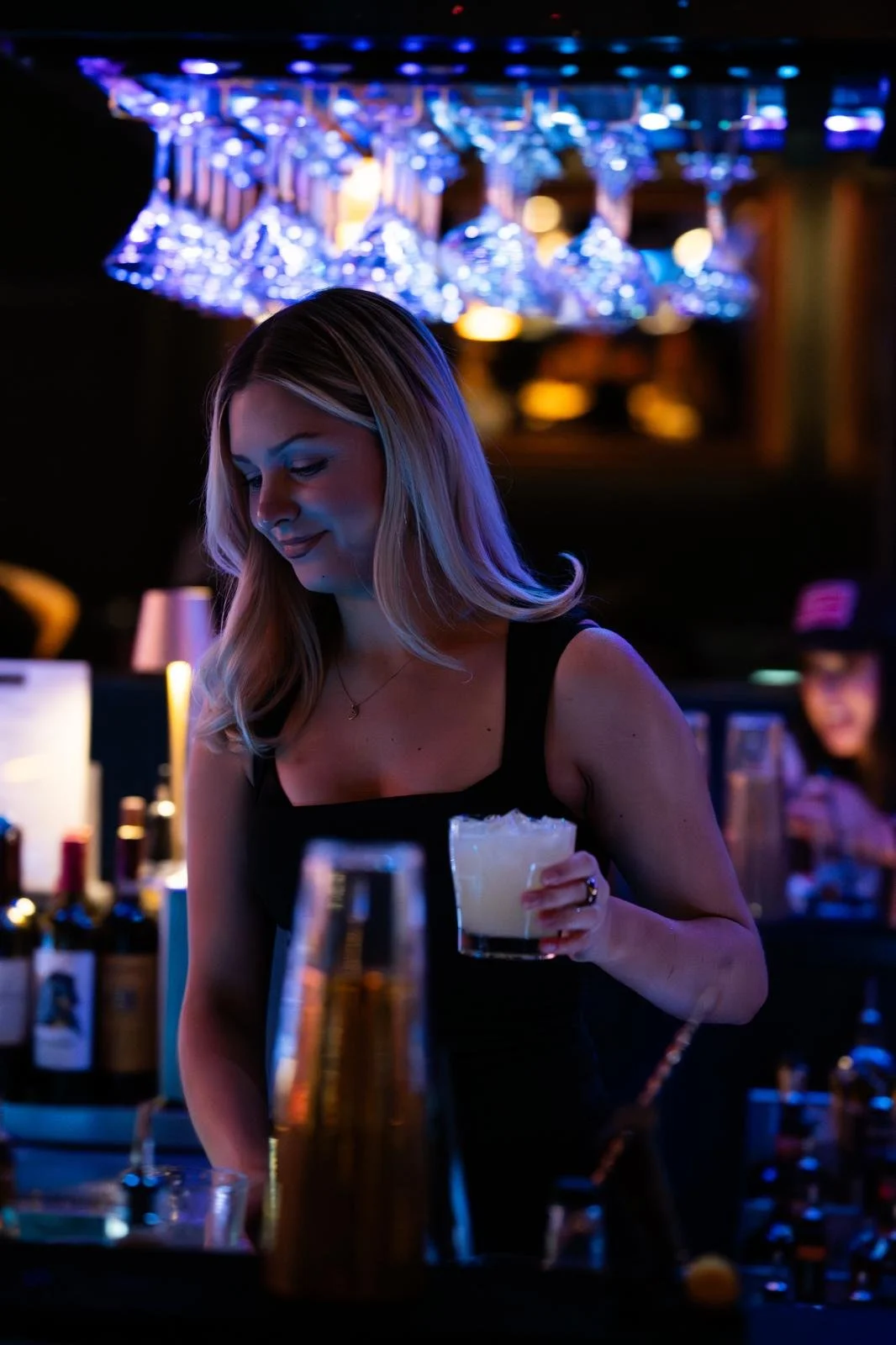 A woman with long blonde hair smiling and holding a drink in a dimly lit bar, with glasses hanging above and other patrons in the background.