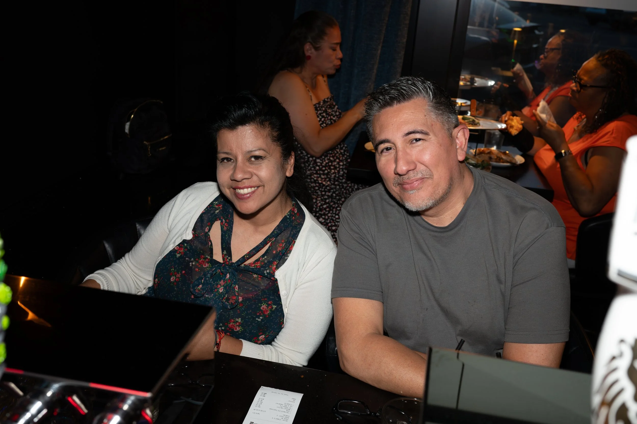 Close-up of smiling woman and man sitting together at a restaurant table, with other diners in the background.