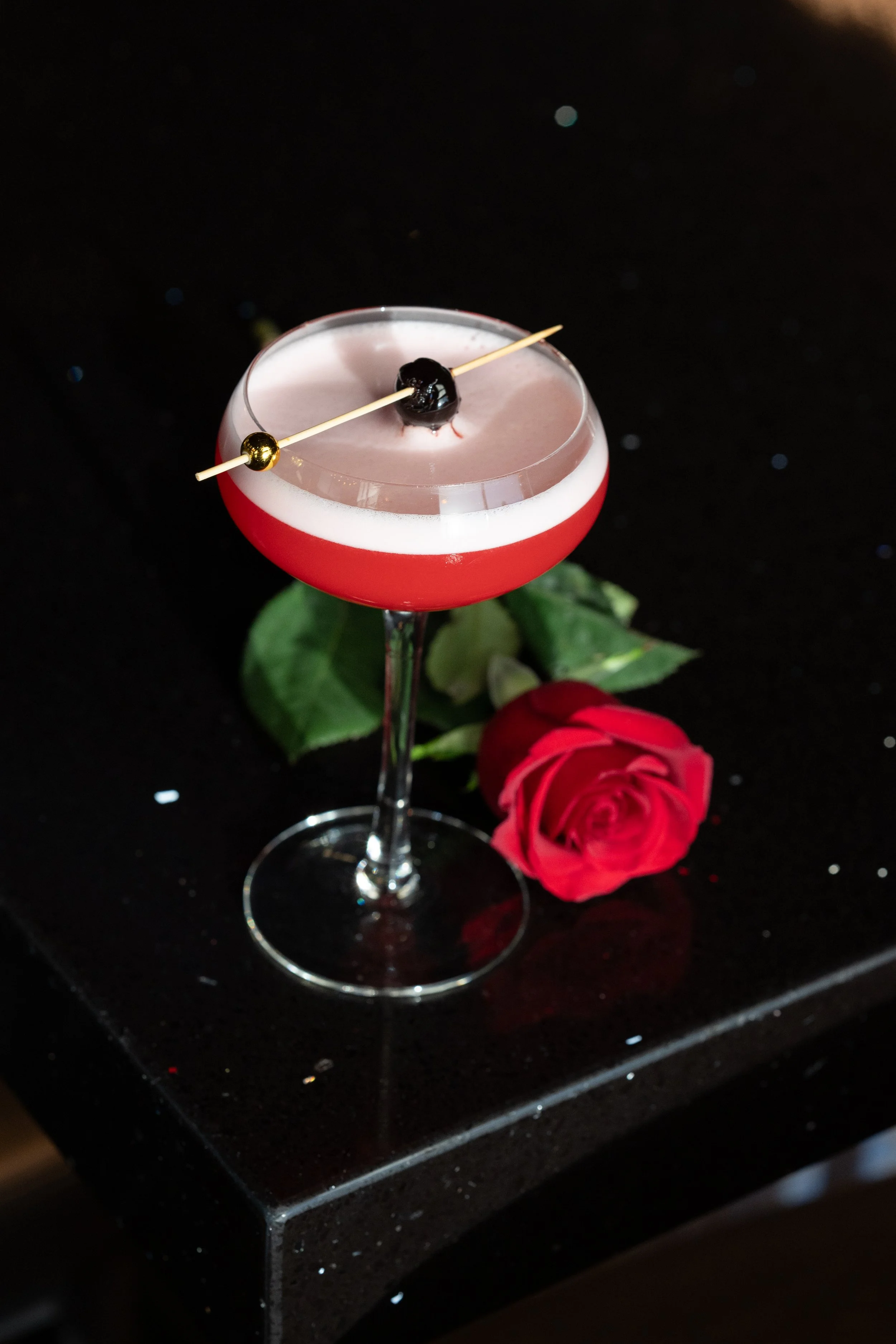 A pink cocktail with a black cherry garnish in a coupe glass, placed on a black surface with a red rose next to it.