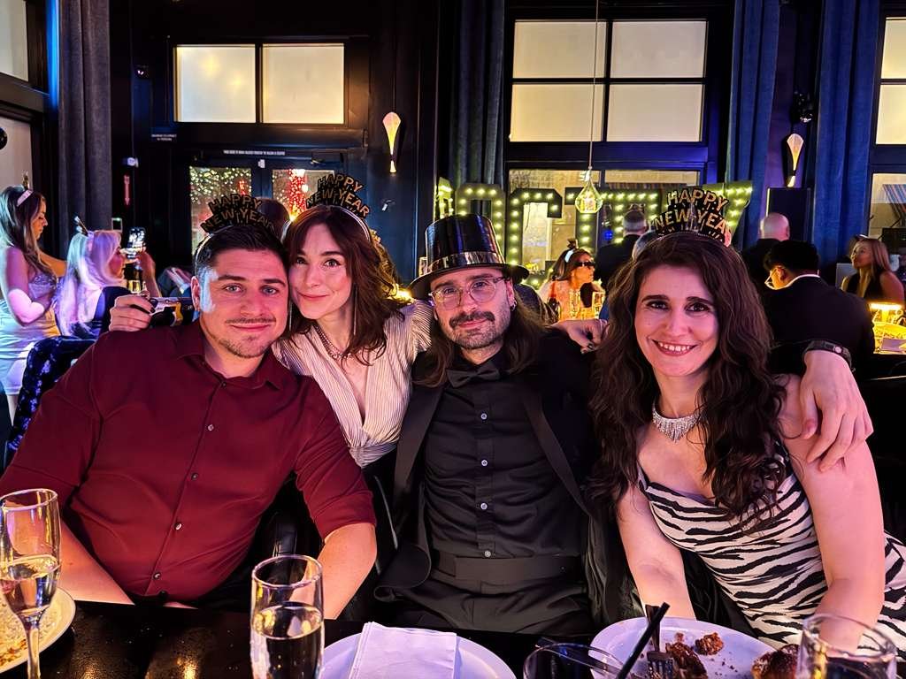 Group of four people celebrating New Year's at a party, wearing festive hats, with a 'Happy New Year' sign and illuminated letters in the background.
