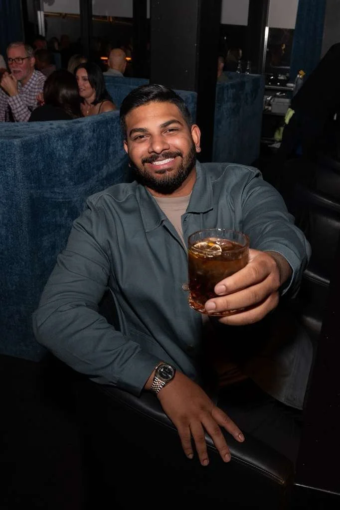 A smiling man with dark hair, beard, and mustache, wearing a gray jacket and watch, holding a glass of dark soda with ice, sitting in a dimly lit restaurant or bar with other patrons in the background.