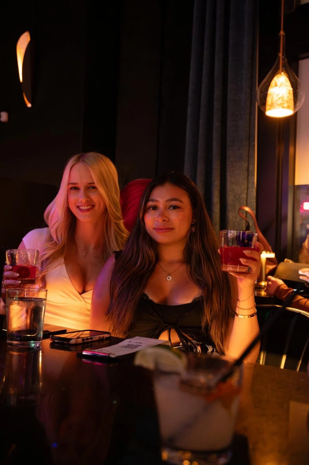 Two women sitting at a restaurant or bar, holding drinks and smiling, with a dessert on the table, dim lighting, and decorative pendant lights in the background.