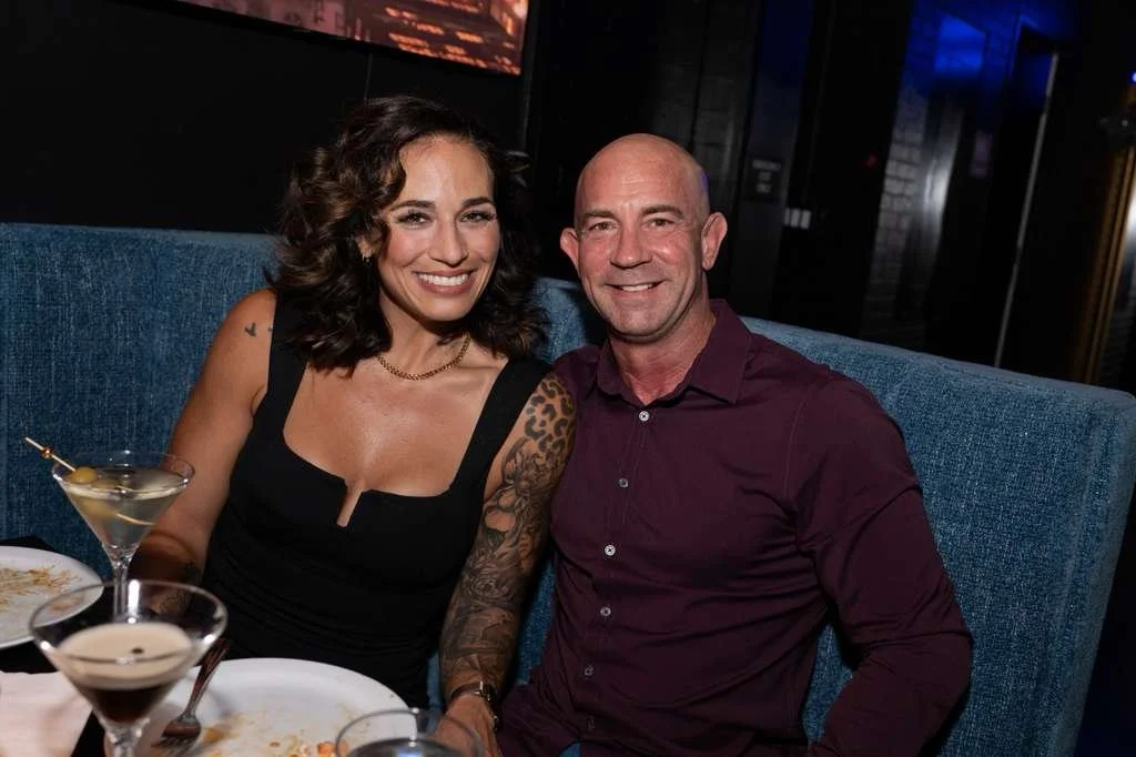 A smiling woman with brown hair and a tattoo on her arm, wearing a black dress, sitting next to a man with a shaved head, wearing a dark red shirt, in a dimly lit restaurant or bar.