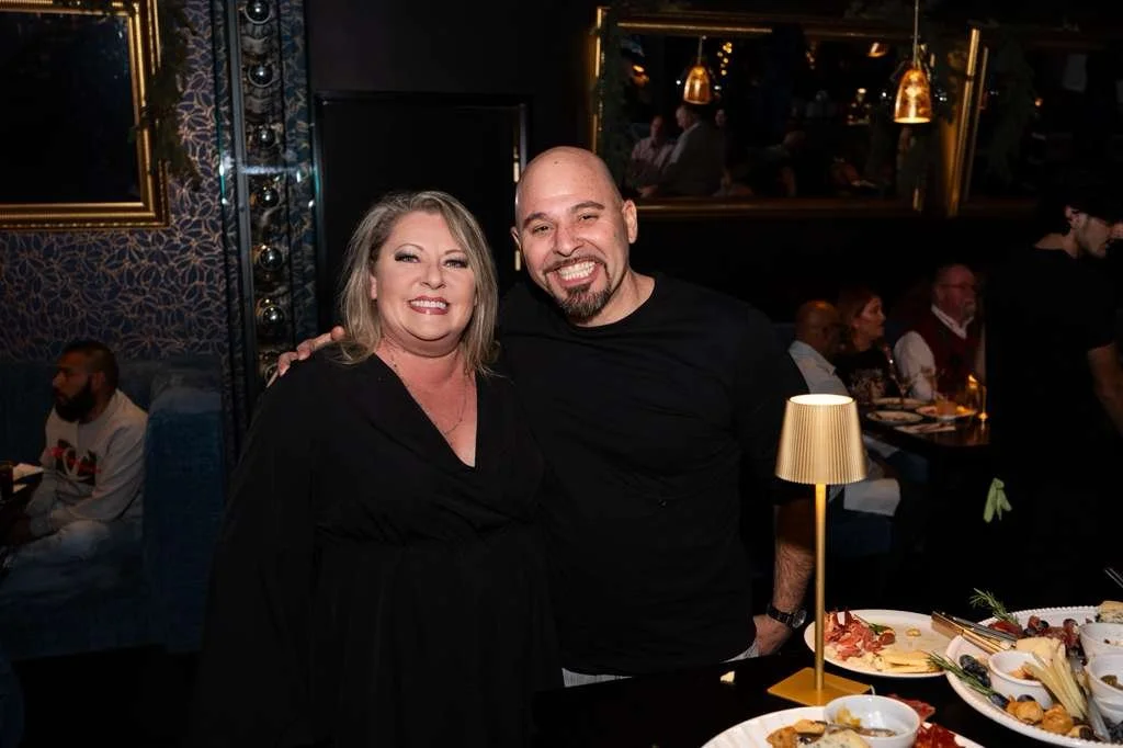 Two adults smiling at a restaurant, with other guests dining in the background, and a table with food and a small lamp in the foreground.
