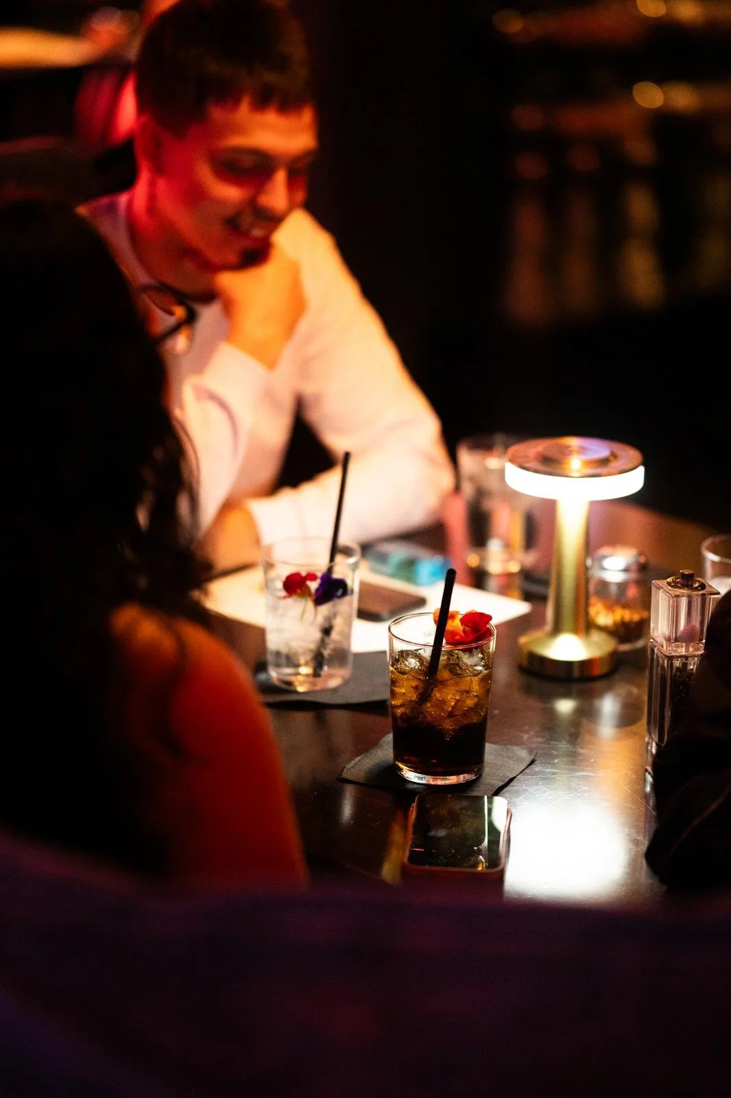 A person is sitting at a dimly lit bar table, smiling, with various drinks and a smartphone on the table, illuminated by a small lamp.