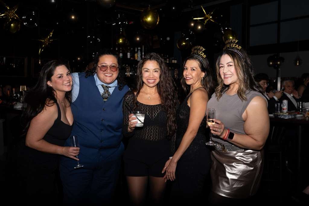 Group of five women celebrating New Year's Eve at a party, wearing 'Happy New Year' hats, holding drinks, in a dimly lit decorated venue.