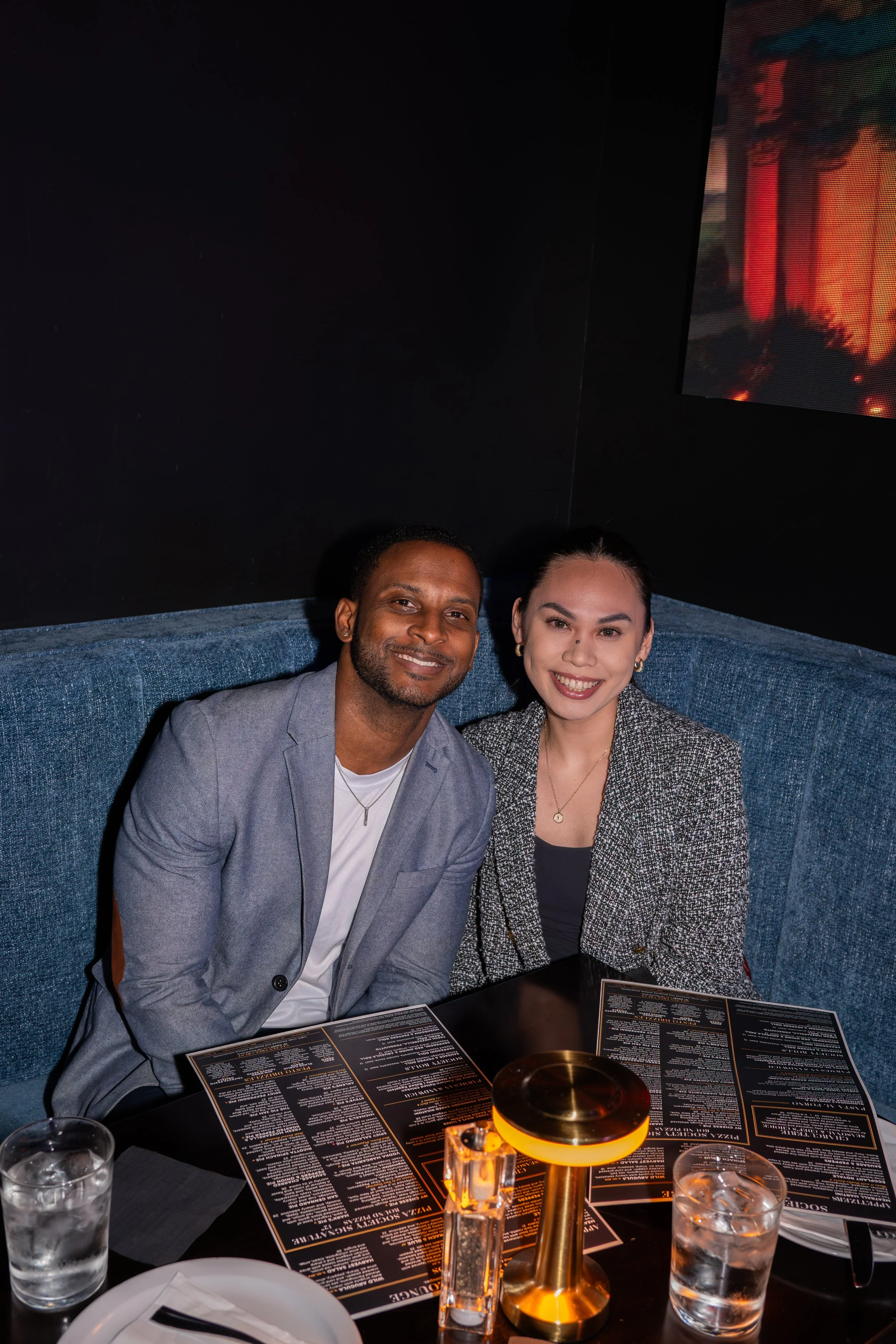 Two people, a man and a woman, smiling at a restaurant table with menus, drinks, and a gold-colored table lamp.