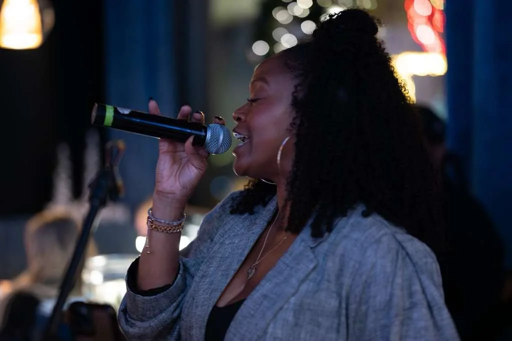 A woman singing into a microphone at what appears to be a live music venue or bar, with holiday lights in the background.