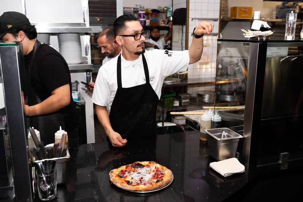 A chef in a white shirt and black apron tosses pizza dough in the kitchen of a restaurant. Several chefs are working in the background, and a freshly baked pizza with toppings is on the counter in front of him.