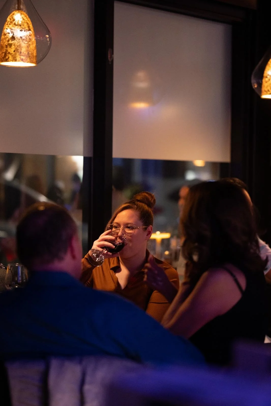 People socializing in a dimly lit restaurant, with a woman wearing glasses and a brown shirt sipping red wine.