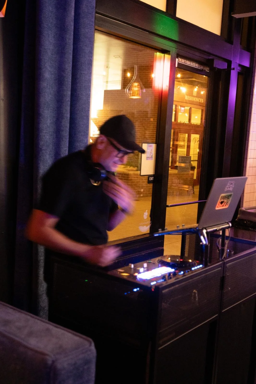 A DJ wearing glasses and a black cap stands at a DJ booth inside a dimly lit venue, with a window showing a brick building outside.