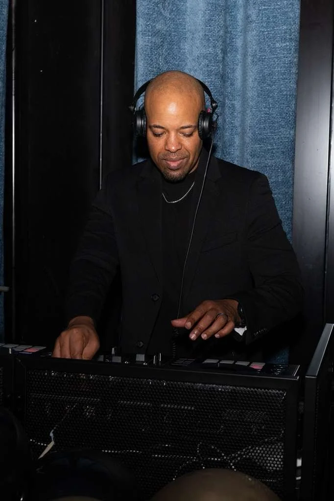 A man with a shaved head wearing headphones and a black suit, standing behind DJ equipment, in a dark setting with black walls and blue fabric panels.