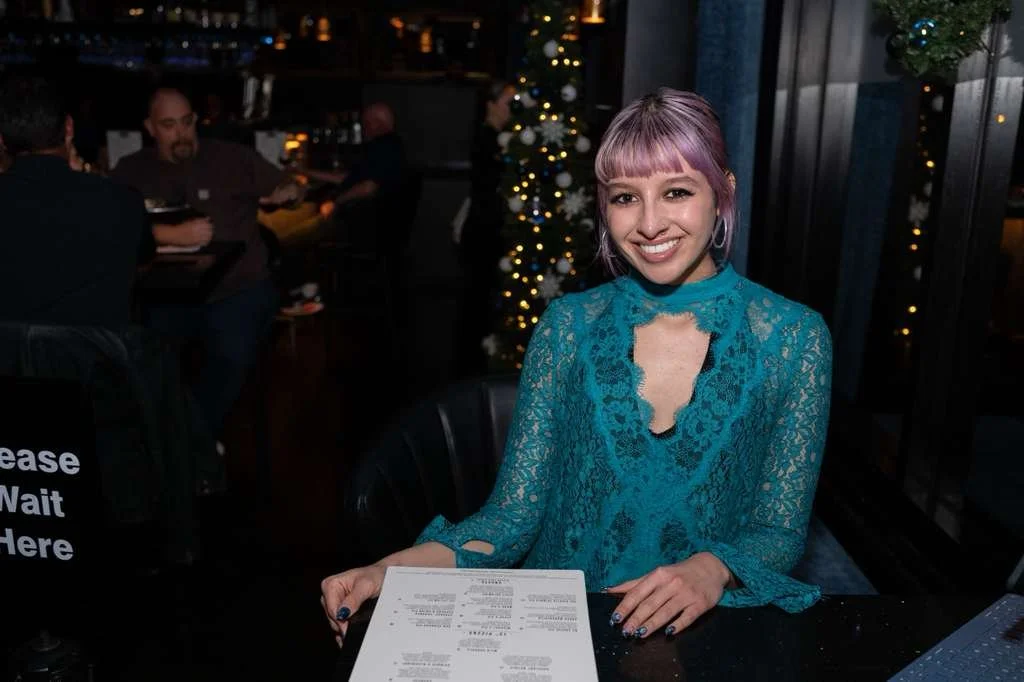 A smiling woman with purple hair and earrings sitting at a table in a restaurant with Christmas lights and a decorated tree in the background.