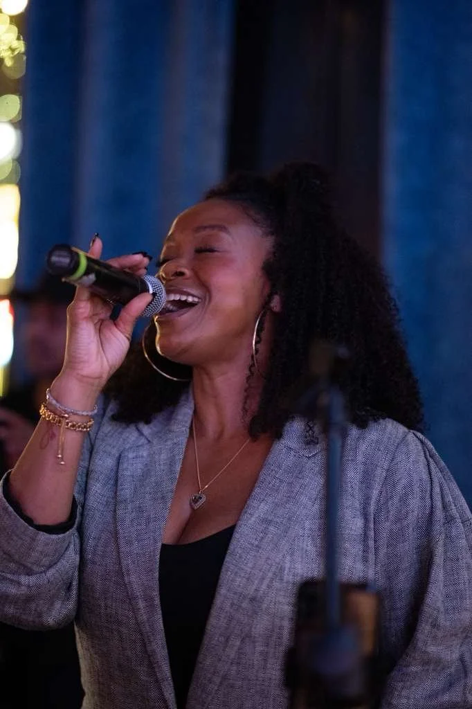 Woman singing into a microphone with eyes closed and smiling, in a likely live performance setting.