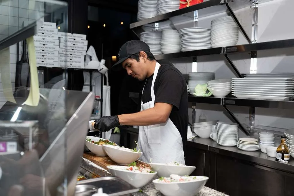 A young man in a black T-shirt, black cap, white apron, and black gloves preparing salads in a commercial kitchen with stacked white plates on shelves behind him.