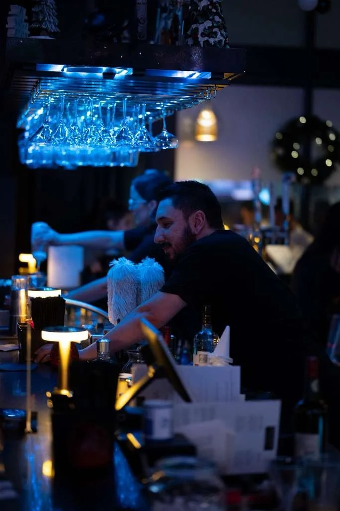 A bartender with short dark hair and a beard working behind a bar counter at night, with hanging upside-down glasses above him, other patrons and bar elements in the background.