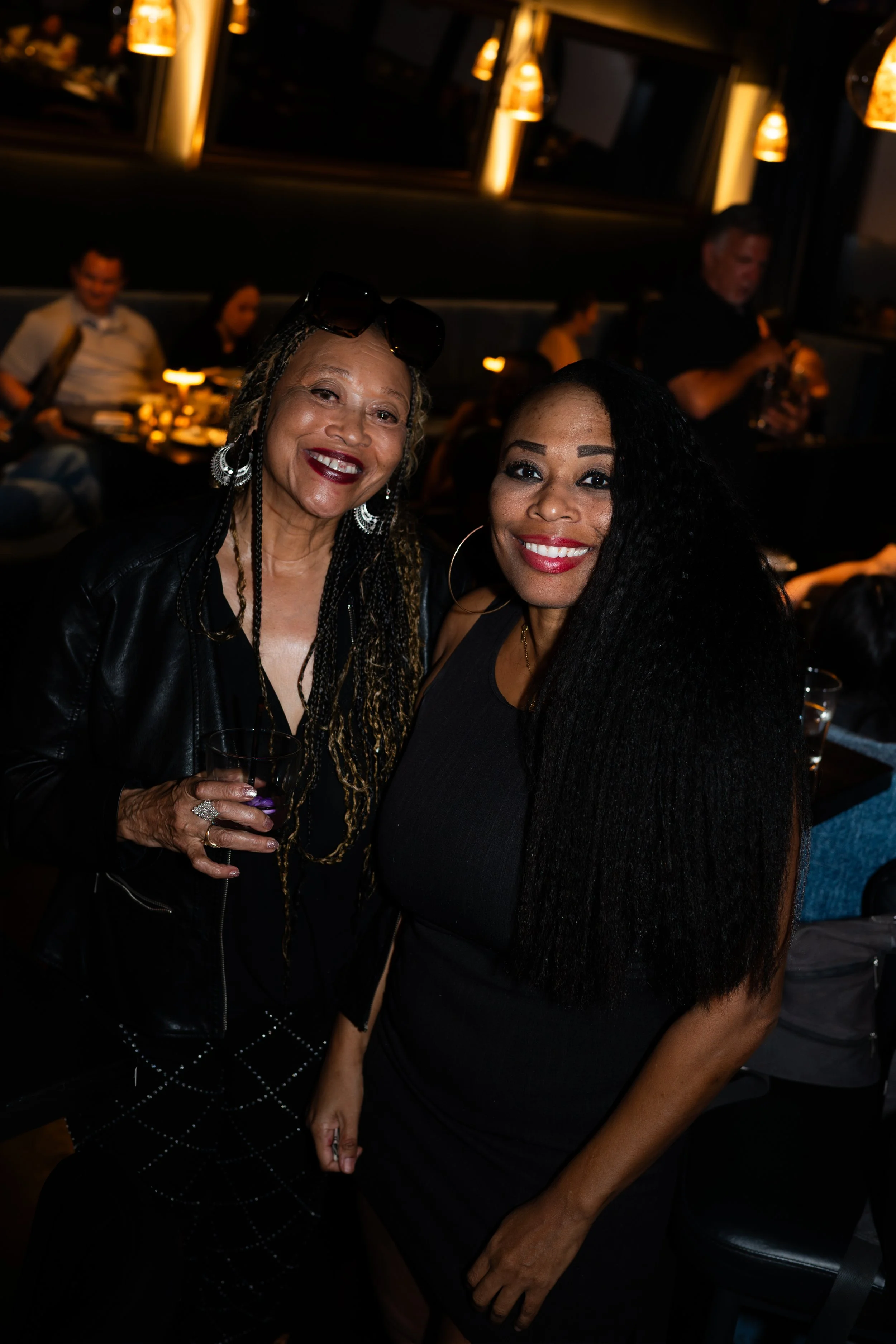 Two women smiling at a social event, with a background of diners and warm lighting.