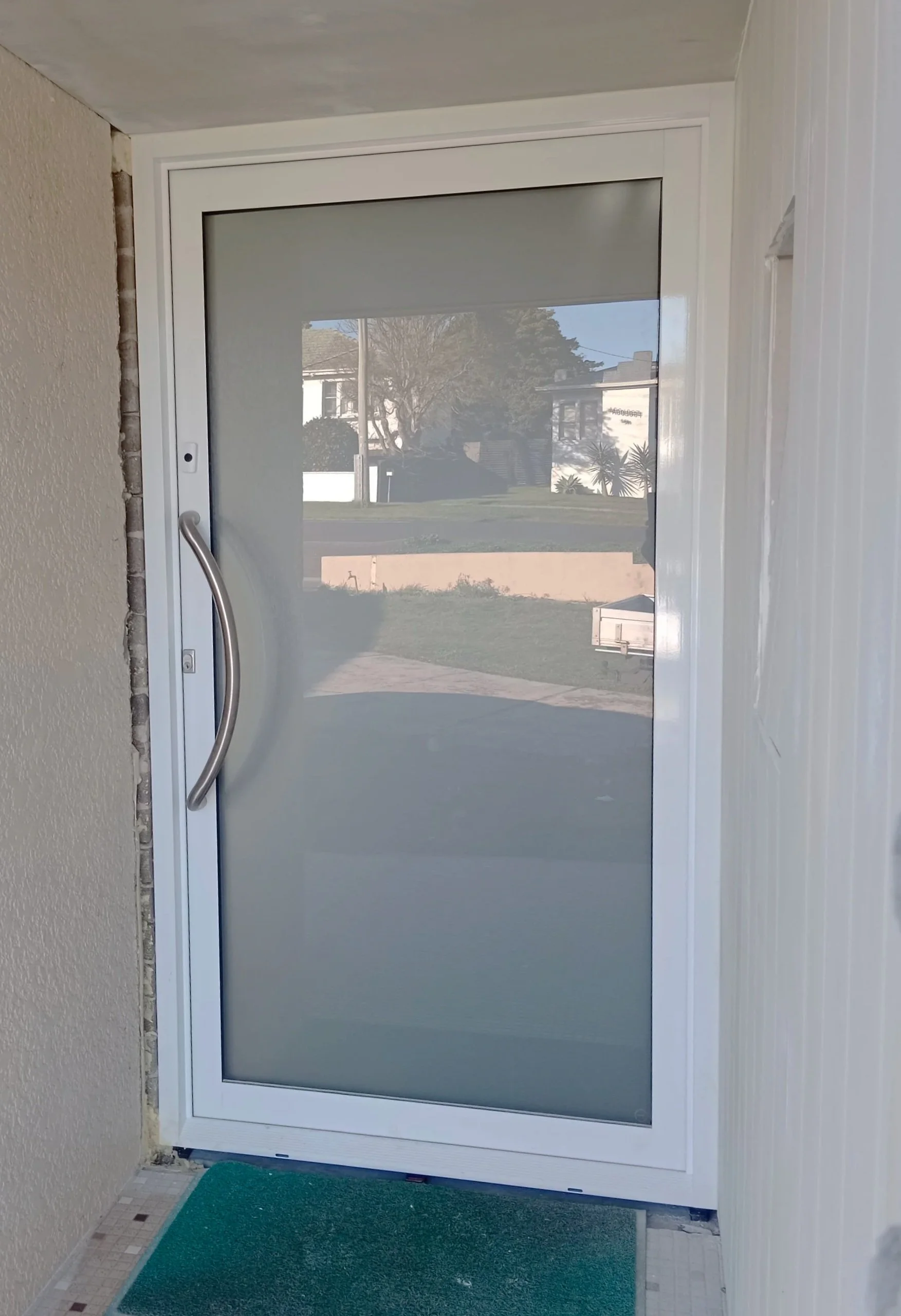 White-framed glass sliding door with a curved metal handle, leading outside to a grassy yard with trees and houses in the background.
