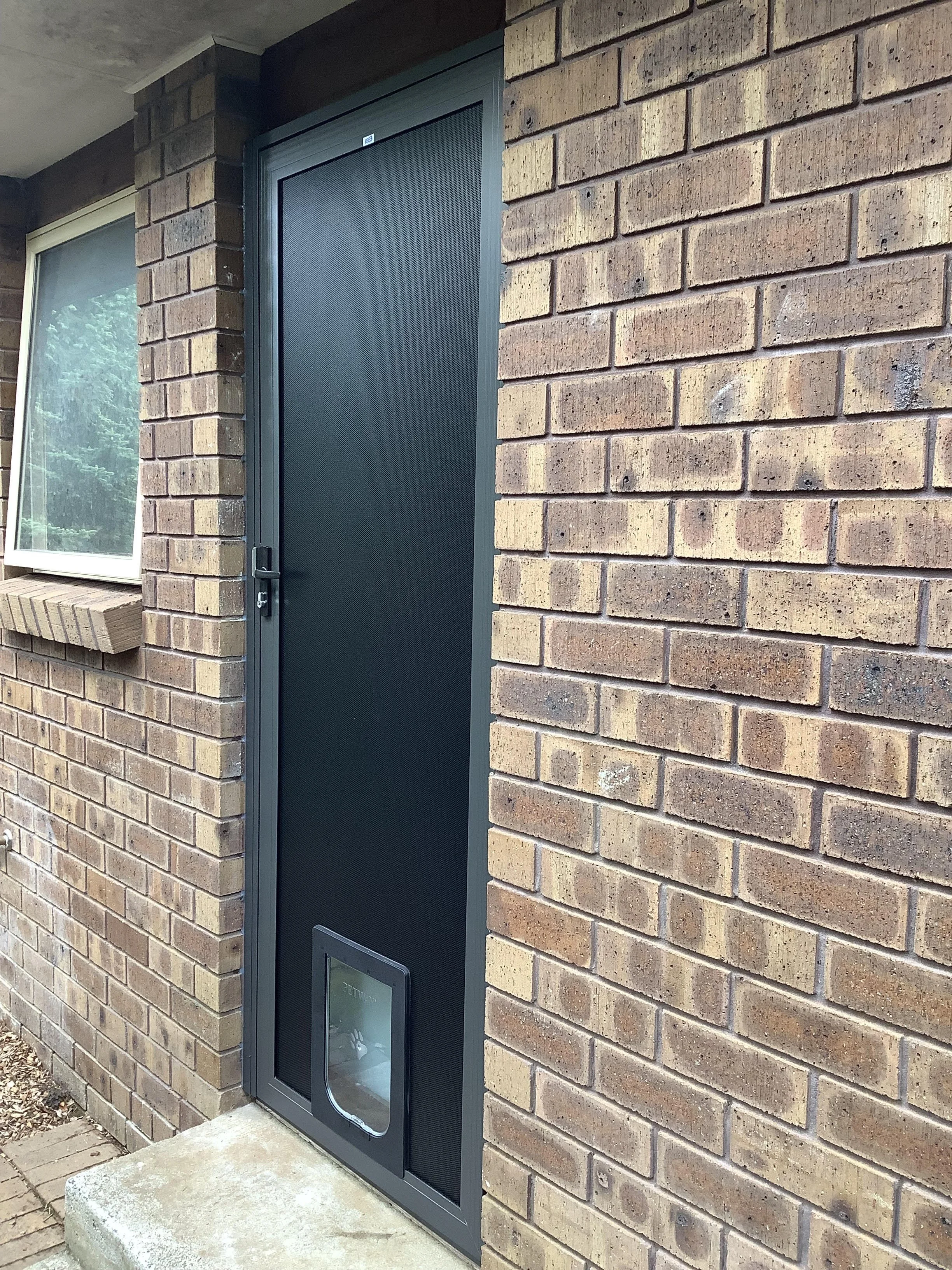Black pet door installed in a black screen door of a brick house.