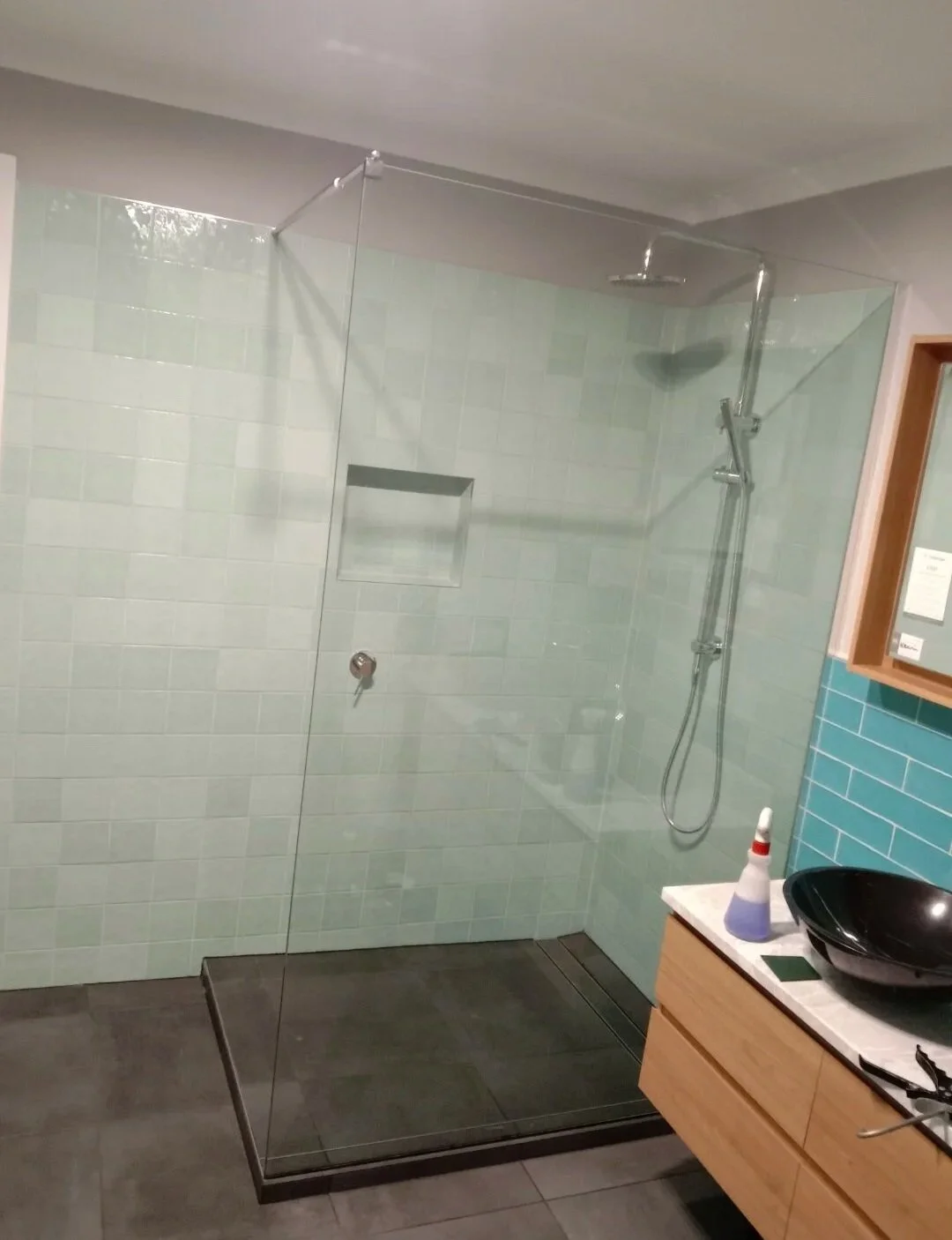 Modern walk-in shower with light green tiled wall, glass enclosure, and built-in niche, adjacent to a wooden vanity with a black vessel sink and a blue spray bottle.