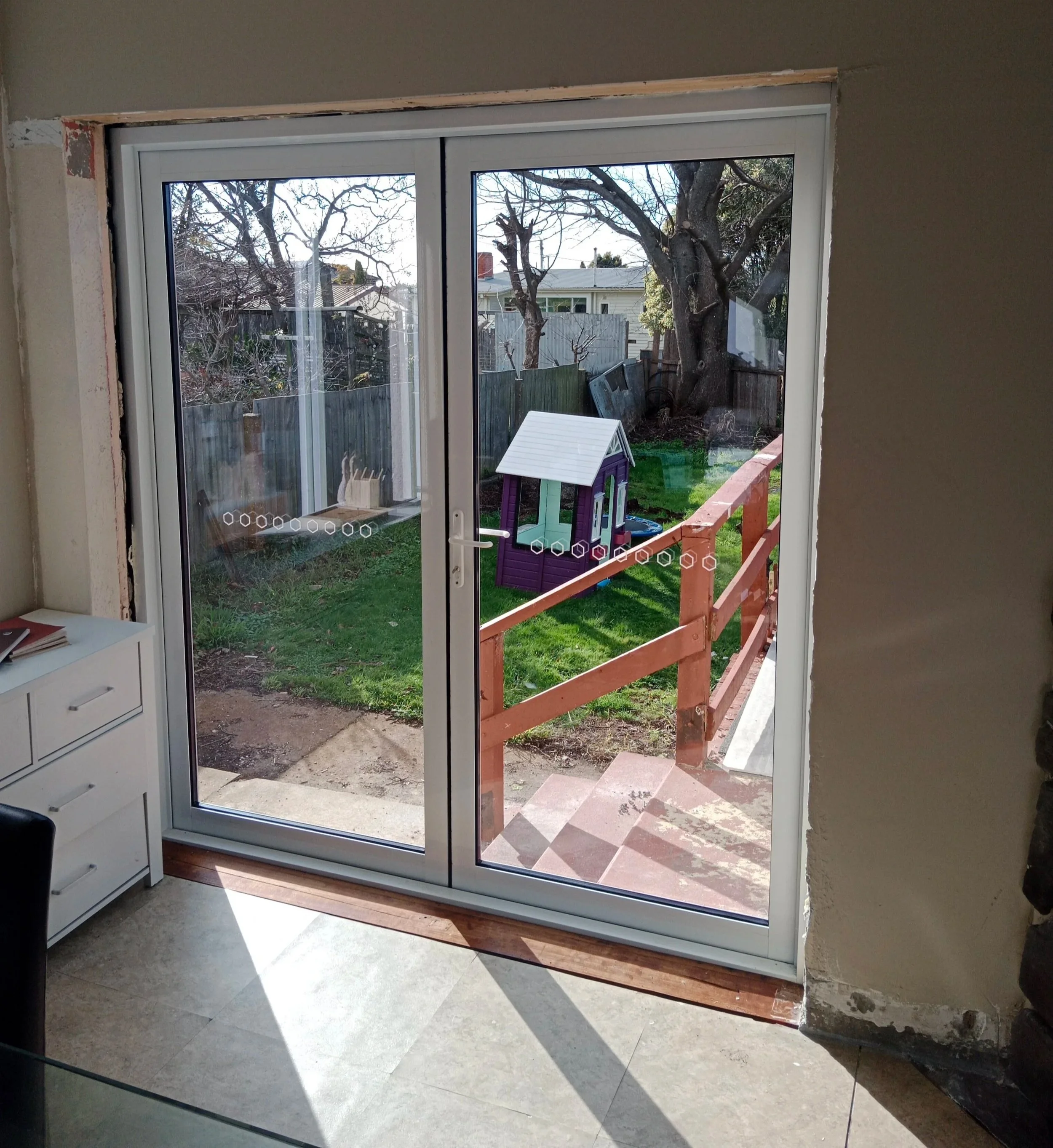 View of a backyard through a sliding glass door, which leads to a wooden deck with stairs, a small purple and white playhouse, green grass, trees, and a wooden fence.