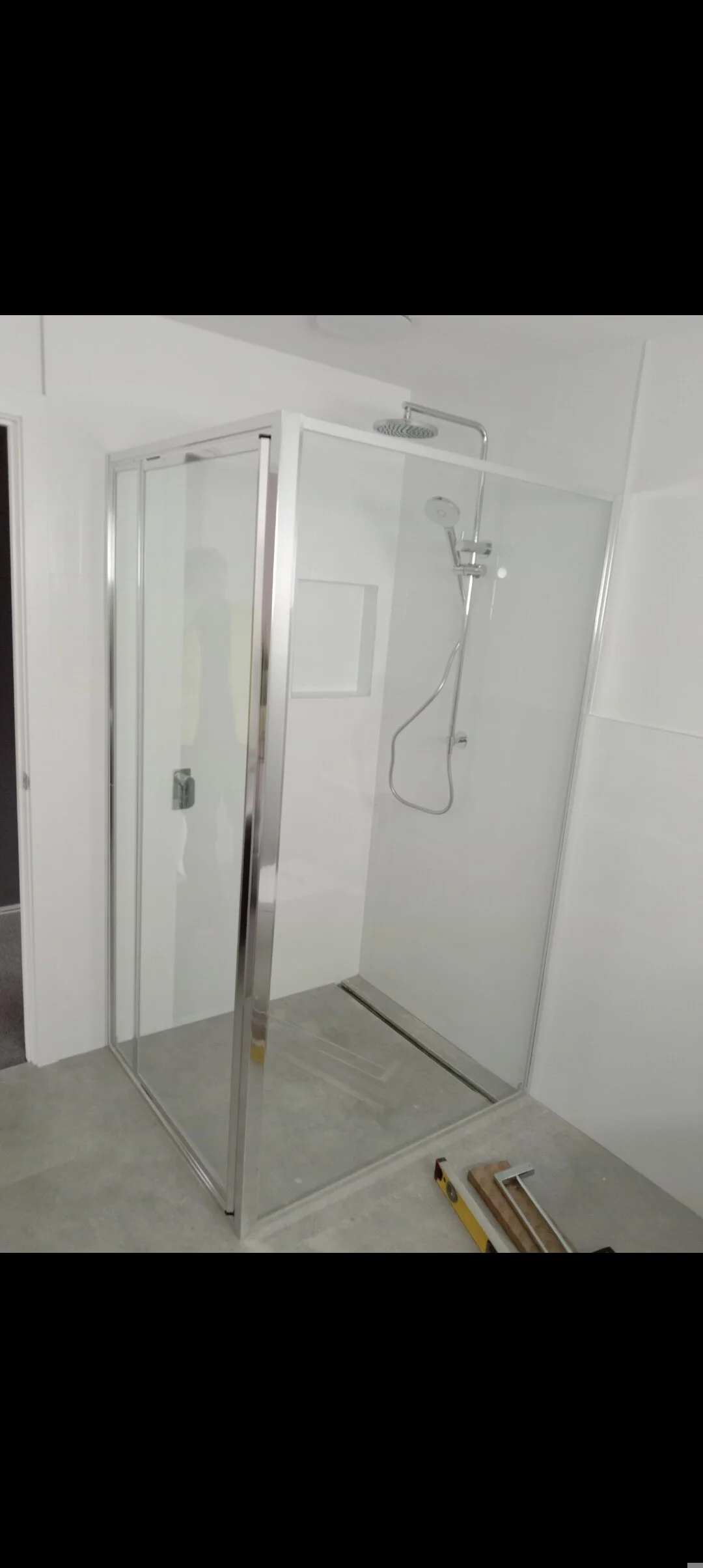 A modern shower enclosure with glass walls and metal framing, featuring a rainfall showerhead and handheld showerhead mounted on the white wall inside. The shower area is unfurnished and appears to be under construction or renovation with tools on the floor.